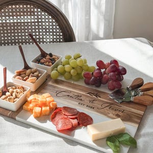 May include: A white and brown rectangular serving board with a marble top and a wooden bottom. The board is engraved with the text "Caitlin & Trevor EST. 2024". The board is filled with various snacks, including grapes, cheese, pepperoni, and nuts.