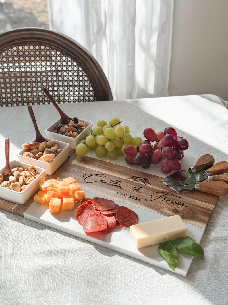 May include: A white and brown rectangular serving board with a marble top and a wooden bottom. The board is engraved with the text "Caitlin & Trevor EST. 2024". The board is filled with various snacks, including grapes, cheese, pepperoni, and nuts.