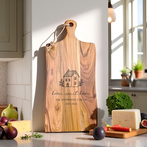 Pu&ograve; includere: Un tagliere rettangolare in legno con manico e corda per appenderlo. Il tagliere presenta una grafica nera di una casa, il testo "home sweet home", "224 DOGWOOD CIRCLE" e "EST. 2025" stampati sulla superficie in venature di legno naturale.