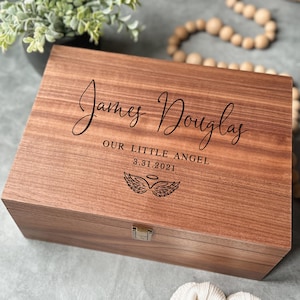 May include: A wooden keepsake box with a hinged lid. The box is engraved with the name "James Douglas", the phrase "OUR LITTLE ANGEL", the date "3.31.2021", and a wing and halo design. The box has a metal clasp.