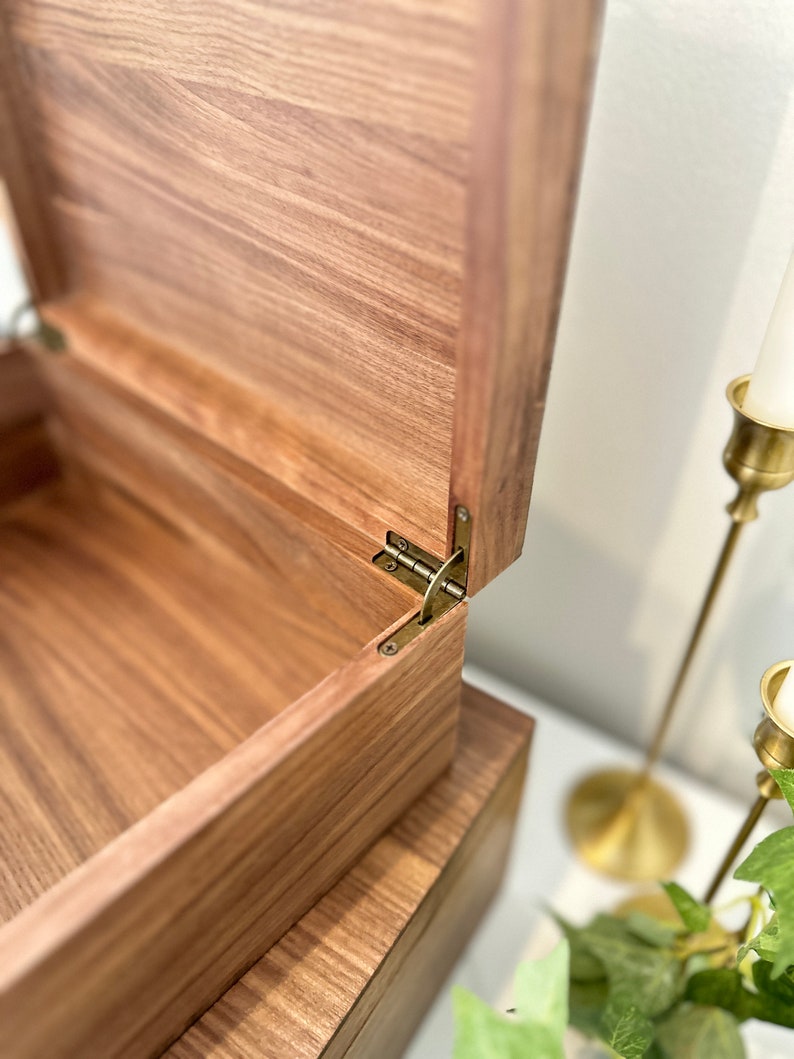 May include: A wooden box with a hinged lid, the lid is open revealing the inside of the box. The box is made of a dark wood with a natural grain pattern. The hinges are made of brass.