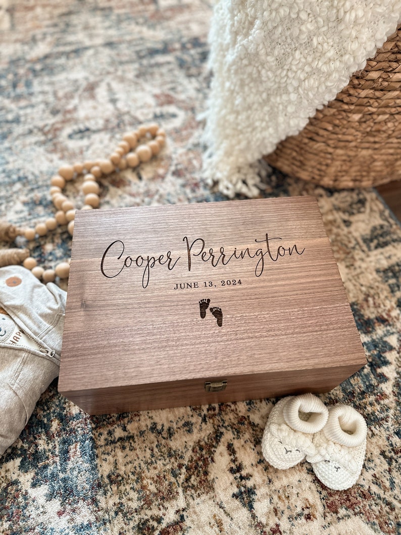 May include: A wooden keepsake box with the inscription "Cooper Perrington June 13, 2024" and a pair of footprints. The box is sitting on a patterned rug.