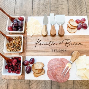 May include: A wooden and marble serving board with a personalised inscription "Kristie + Brian EST. 2024". The board is filled with various snacks, including grapes, cheese, crackers, salami, and nuts.