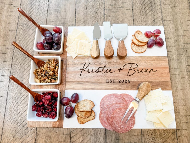 May include: A wooden and marble serving board with a personalised inscription "Kristie + Brian EST. 2024". The board is filled with various snacks, including grapes, cheese, crackers, salami, and nuts.