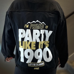May include: Black denim jacket with a white and gold graphic that says "I'm Primed to Party Like It's 1990" and "Let's Go Colorado!" with a logo that says "Slick" underneath.