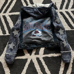 May include: A blue denim jacket with a gray hooded sweatshirt attached. The jacket has a Colorado Avalanche logo on the back with the words "Colorado Avalanche". The sleeves have silver glitter stars.