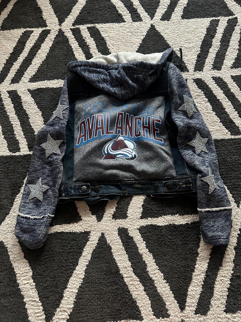 May include: A blue denim jacket with a gray hooded sweatshirt attached. The jacket has a Colorado Avalanche logo on the back with the words "Colorado Avalanche". The sleeves have silver glitter stars.