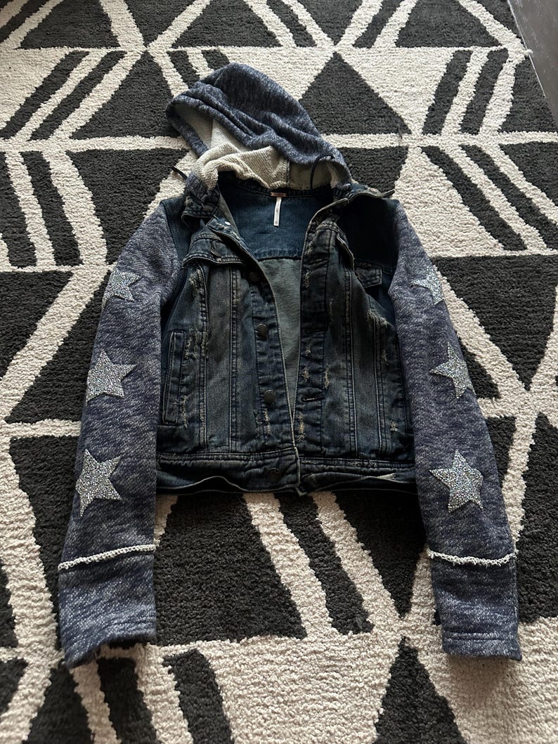 May include: A blue denim jacket with a hooded sweatshirt attached to the sleeves. The sleeves have silver glitter stars on them.