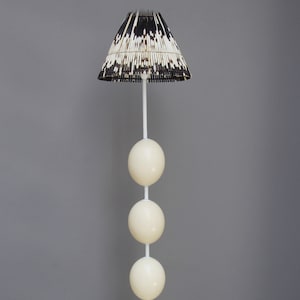 Può includere: Una lampada da terra bianca con un paralume unico realizzato con materiali naturali intrecciati. Il paralume ha un motivo bianco e nero. La lampada ha una serie di cinque sfere bianche attaccate al palo.