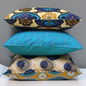 May include: A stack of three decorative throw pillows with colourful geometric and floral patterns. The top pillow is yellow, blue and black with a floral pattern. The middle pillow is turquoise blue with a wavy pattern. The bottom pillow is yellow, blue and black with a circular pattern.