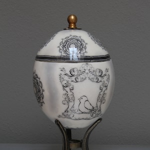 May include: A white egg with black and gold decorative designs, including a bird, is displayed on a gold metal stand.