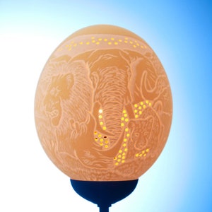 Könnte beinhalten: Eine geschnitzte Straußeneierlampe mit einem Design aus Elefant, Löwe und anderen Tieren. Die Lampe ist beleuchtet und das Licht scheint durch die Löcher im Ei.