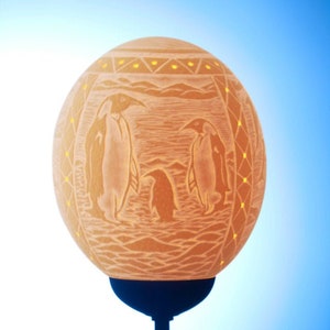 Könnte beinhalten: Eine geschnitzte Straußeneierlampe mit einem Design aus drei Pinguinen. Die Lampe ist eingeschaltet und das Licht scheint durch die Schnitzereien. Die Lampe steht auf einem schwarzen Sockel.