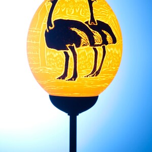 May include: A yellow ostrich egg lamp with a black silhouette of three ostriches carved into the surface. The lamp is on a black stand.