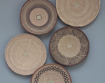 Könnte beinhalten: Fünf runde Wandkörbe aus geflochtenem Material in verschiedenen Naturtönen und Mustern. Jeder Korb hat ein einzigartiges Design, darunter Spiral-, geometrische und strukturierte Muster. Die Körbe werden vor einem grauen Hintergrund präsentiert.