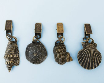 May include: Four gold-toned curtain tiebacks with different seashell designs. The tiebacks feature a clip at the top and a seashell design at the bottom. The seashells include a conch shell, a sand dollar, a scallop shell, and a clam shell.