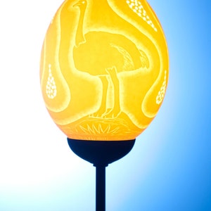 Può includere: Una lampada a forma di uovo di struzzo giallo con un design intagliato di uno struzzo. La lampada è su un piedistallo nero.