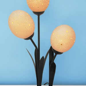 May include: A black metal table lamp with three ostrich egg shades. Each shade has a white starburst pattern and is illuminated from within.