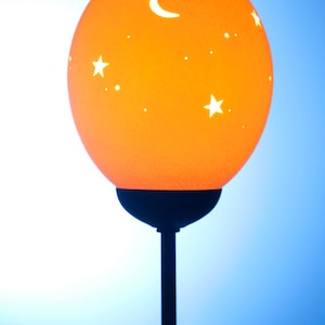 Könnte beinhalten: Eine Lampe mit einem orangefarbenen, kugelförmigen Schirm, der eine sichelförmige Mondsichel und Sterne ausschneidet. Der Schirm sitzt auf einem schwarzen Metallständer.