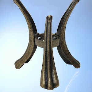 Può includere: Un supporto decorativo in metallo dorato con una parte superiore rotonda e tre gambe. Le gambe sono curve e hanno un design decorativo.