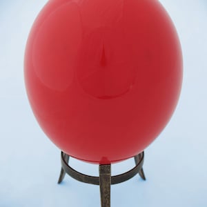 Könnte beinhalten: Eine leuchtend rote Glaskugel auf einem schwarzen Metallständer mit drei Beinen.