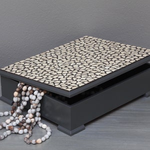 May include: A gray wooden box with a mosaic tile lid. The lid is open and a strand of white and brown beads is spilling out.