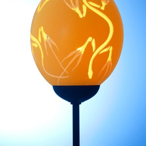 Könnte beinhalten: Eine dekorative Lampe aus einem Straußenei mit aufwendigen Schnitzereien. Das Ei ist hellorange und die Schnitzereien sind dunkelorange. Die Lampe steht auf einem schwarzen Sockel.