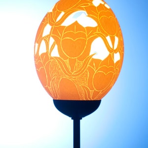Könnte beinhalten: Eine Lampe aus einem geschnitzten Straußenei mit floralem Design. Das Ei ist hellorange und auf einem schwarzen Metallständer montiert.