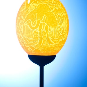 Könnte beinhalten: Eine gelbe Straußen-Eierlampe mit einem geschnitzten Design aus Straußen und Blättern. Die Lampe steht auf einem schwarzen Ständer.