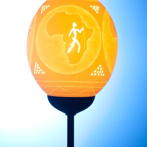 Könnte beinhalten: Eine geschnitzte Straußeneierlampe mit einer Silhouette des afrikanischen Kontinents und einer Person, die mit einem Speer geht. Die Lampe steht auf einem schwarzen Sockel vor blauem Hintergrund.
