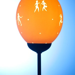 Könnte beinhalten: Eine Lampe mit einem orangefarbenen, kugelförmigen Schirm, der weiße Silhouetten von Menschen zeigt. Die Lampe hat einen schwarzen Metallfuß und einen Ständer.