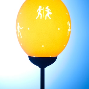 Könnte beinhalten: Eine gelbe Lampe mit einem runden Schirm, der weiße Silhouetten von tanzenden Menschen zeigt. Die Lampe hat einen schwarzen Metallfuß und einen Stiel.