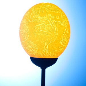 May include: A yellow lampshade with an intricate carved design of birds and a house. The lampshade is on a black metal stand.