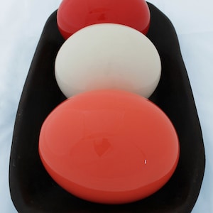 May include: Three glossy spheres, one red, one white, and one coral, are arranged in a stack on a dark brown wooden tray.