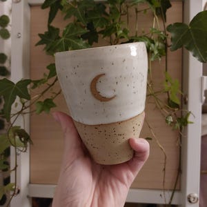 Può includere: Una tazza in ceramica fatta a mano con una parte superiore color crema e una parte inferiore macchiata marrone chiaro. Un design a mezzaluna è centrato sulla sezione color crema. La tazza è tenuta in mano, con viti verdi sullo sfondo.