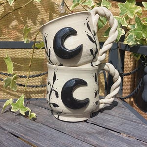 Pode incluir: Duas canecas de cerâmica empilhadas com um esmalte de cor creme, cada uma com um emblema de lua crescente preta em relevo e motivos de videiras botânicas pintados à mão. As canecas têm uma alça distinta em estilo de corda torcida.