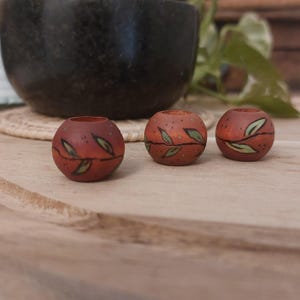 Op de afbeelding: Drie kleine, ronde, terracotta-kleurige kandelaars met groene bladeren. De kandelaars zijn gerangschikt op een houten oppervlak, met een donkere kom en groen in de achtergrond.