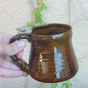 May include: A glossy, dark brown ceramic mug with a wide, rounded body and a thick, twisted handle. A small, flat, light-colored decorative element shaped like a leaf or petal is attached to the top of the handle where it meets the mug rim.