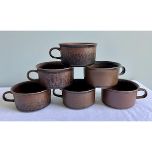 May include: A set of six brown ceramic soup mugs arranged in a pyramid stack. Each mug features a wide, cylindrical body with a single side handle and a mottled, dark-to-light brown glaze finish that varies in texture across the surface of each piece.