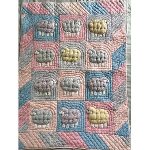 May include: A rectangular quilted blanket featuring a grid pattern of twelve individual sheep appliques. The sheep are rendered in shades of gray, yellow, and floral patterns. The background consists of alternating pink and blue striped fabric blocks.
