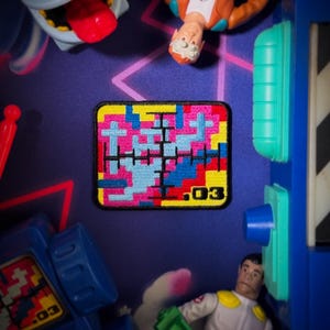 May include: Embroidered patch with a colorful pixelated design in pink, blue, yellow, and black. The patch has the number ".03" in yellow. The patch is surrounded by Ghostbusters action figures and props.