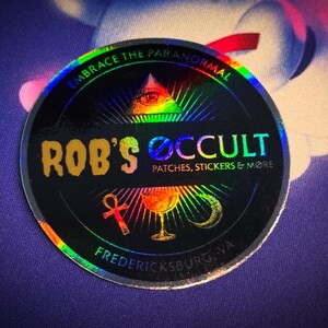 Może przedstawiać: Holograficzna naklejka z tekstem "Rob's Occult" w złotych literach. Naklejka ma czarne tło z tęczową obwódką i przedstawia wszechwidzące oko, symbol ankh i kielich. Tekst "Embrace the Paranormal" jest napisany w tęczy nad logo. Tekst "Patches, Stickers & More" jest napisany pod logo. Tekst "Fredericksburg, VA" jest napisany na dole naklejki.