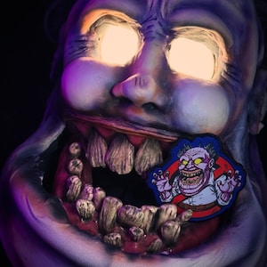 May include: A large, grotesque, purple-toned face with glowing yellow eyes and a wide open mouth filled with large, uneven teeth. A colourful patch featuring a cartoon character with the Ghostbusters logo is placed in the mouth.
