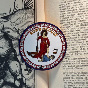 May include: A circular sticker featuring the text ROB'S OCCULT in orange letters. The design shows a person in a red dress standing over a person in a blue shirt, holding a black object, with a coffee pot nearby, all inside a red and white border.