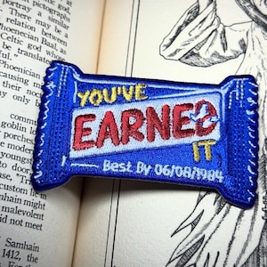 May include: A blue embroidered patch in the shape of a candy bar. The patch reads "YOU'VE EARNED IT" in red and gold thread, with "Best By 06/08/1984" below. The patch is on a book page.