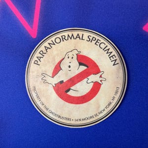 May include: A round, cream-colored coaster with a red "no ghost" symbol and a cartoon ghost inside. The text "PARANORMAL SPECIMEN" is printed in a circle around the symbol. The text "PROPERTY OF THE GHOSTBUSTERS 14 N MOORE ST. NEW YORK, NY 10013" is printed around the bottom of the coaster.