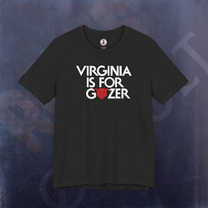 May include: A black short-sleeved t-shirt featuring the white text VIRGINIA IS FOR GOZER with a red graphic of a creature's head replacing the letter O in the word GOZER. The shirt has a round neckline and a small circular logo on the inner collar.