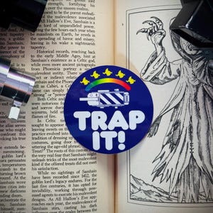May include: A blue and white sticker with a rainbow and a camera design. The sticker says "TRAP IT!" in white text. The sticker is placed on a book with text about the Celtic god Samhain.