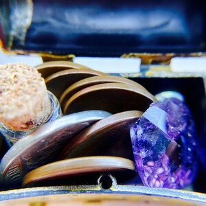 K&ouml;nnte beinhalten: Eine kleine, verzierte Schatztruhe aus Messing ist ge&ouml;ffnet und zeigt einen Stapel Silber- und Kupferm&uuml;nzen, eine kleine Glasflasche mit einem Korkstopfen und einen gro&szlig;en, facettierten, violetten Amethystkristall.