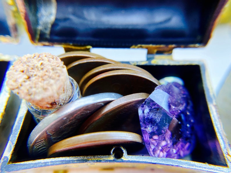 K&ouml;nnte beinhalten: Eine kleine, verzierte Schatztruhe aus Messing ist ge&ouml;ffnet und zeigt einen Stapel Silber- und Kupferm&uuml;nzen, eine kleine Glasflasche mit einem Korkstopfen und einen gro&szlig;en, facettierten, violetten Amethystkristall.